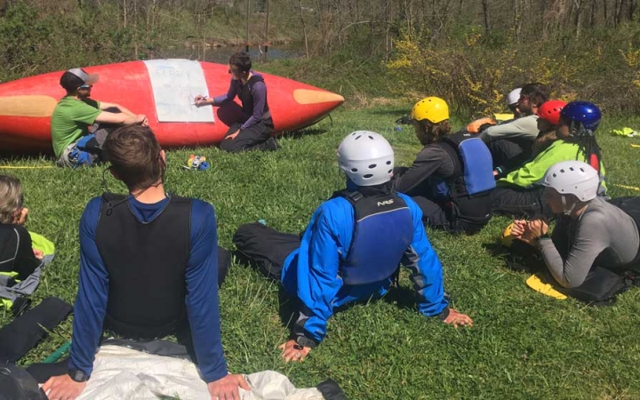 An instructor points to a sheet of paper laid over an upside-down canoe. A group of students rest on the grass, watching.